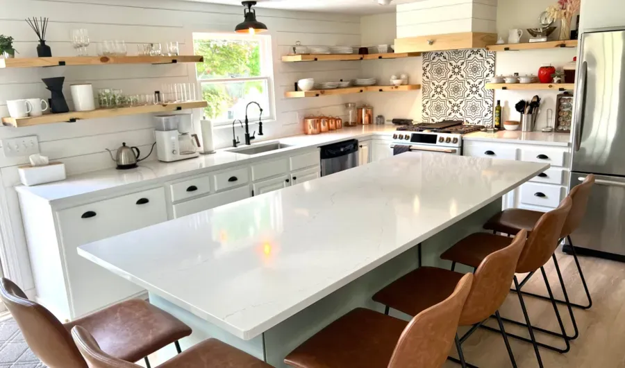 Bright modern kitchen with a large white island, wood chairs, open shelves, and patterned backsplash.