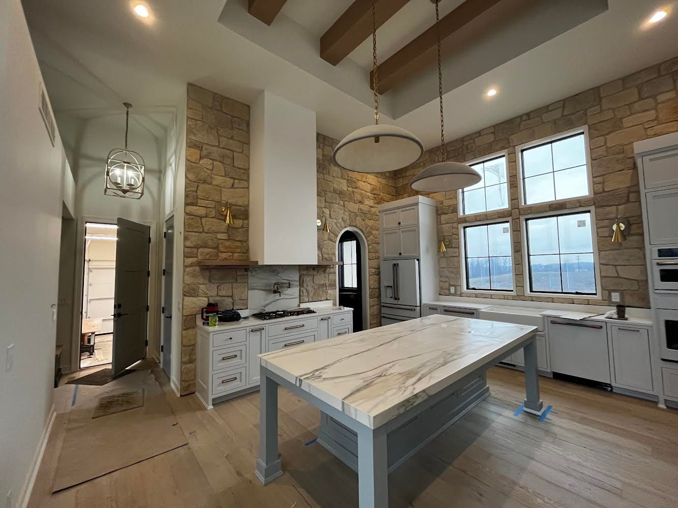 Bright rustic kitchen with stone walls, white cabinets, large windows, and a central island table.