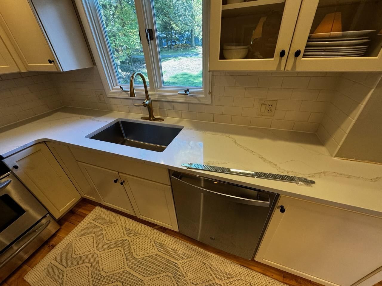Modern kitchen with white countertops, black sink, stainless dishwasher, and window above the sink