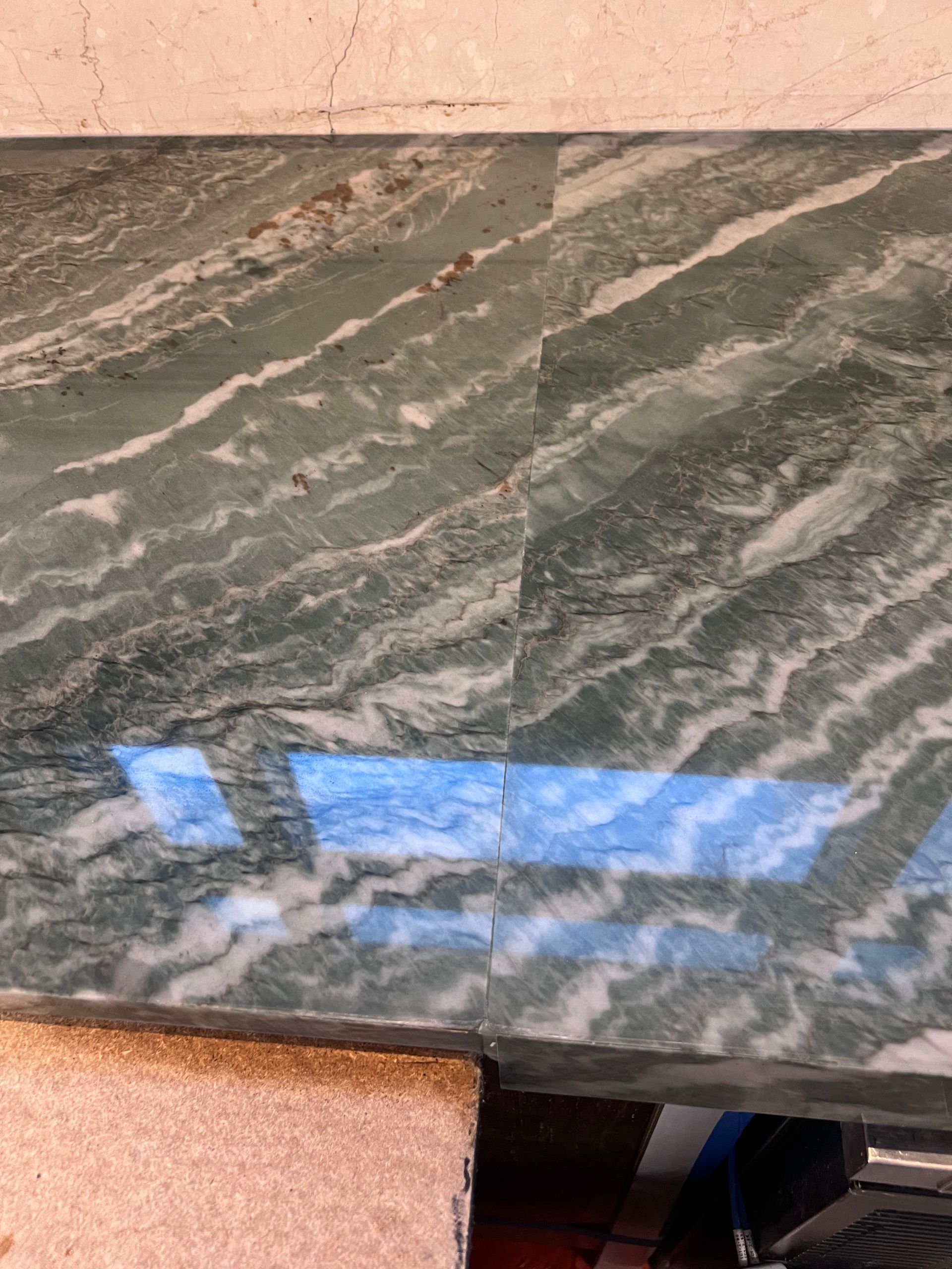 Close-up of a gray-green veined stone countertop with a blue window reflection and a pink edge below