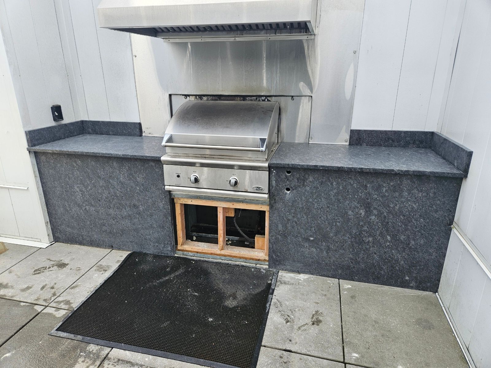 Outdoor built-in stainless steel grill with stone counters and a black mat on concrete patio.