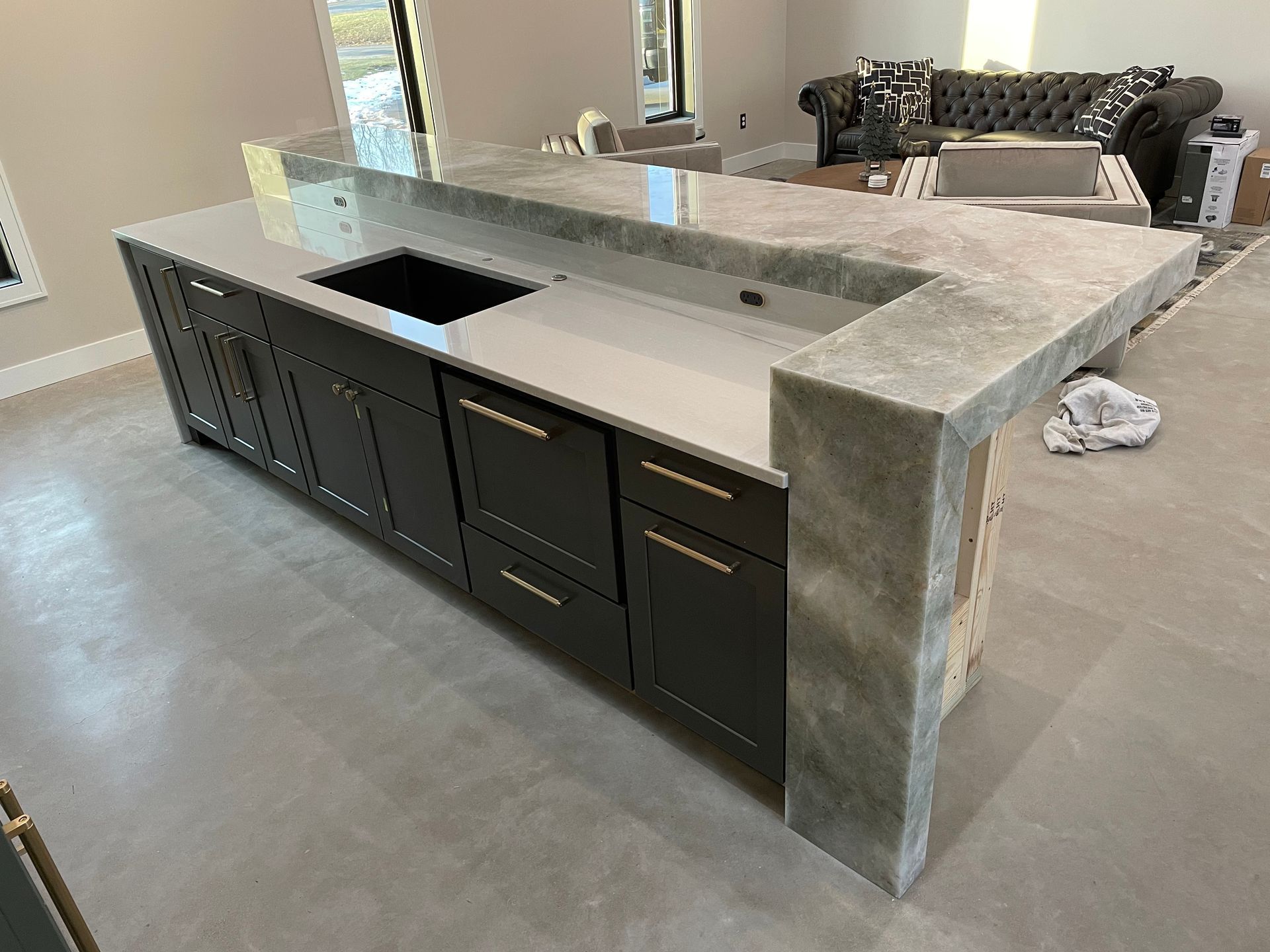 Large modern kitchen island with gray stone countertop, dark cabinets, and built-in sink in a bright room
