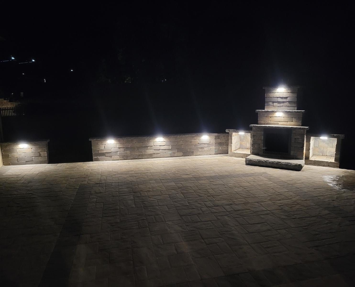 A brick patio with a fireplace and lights at night.