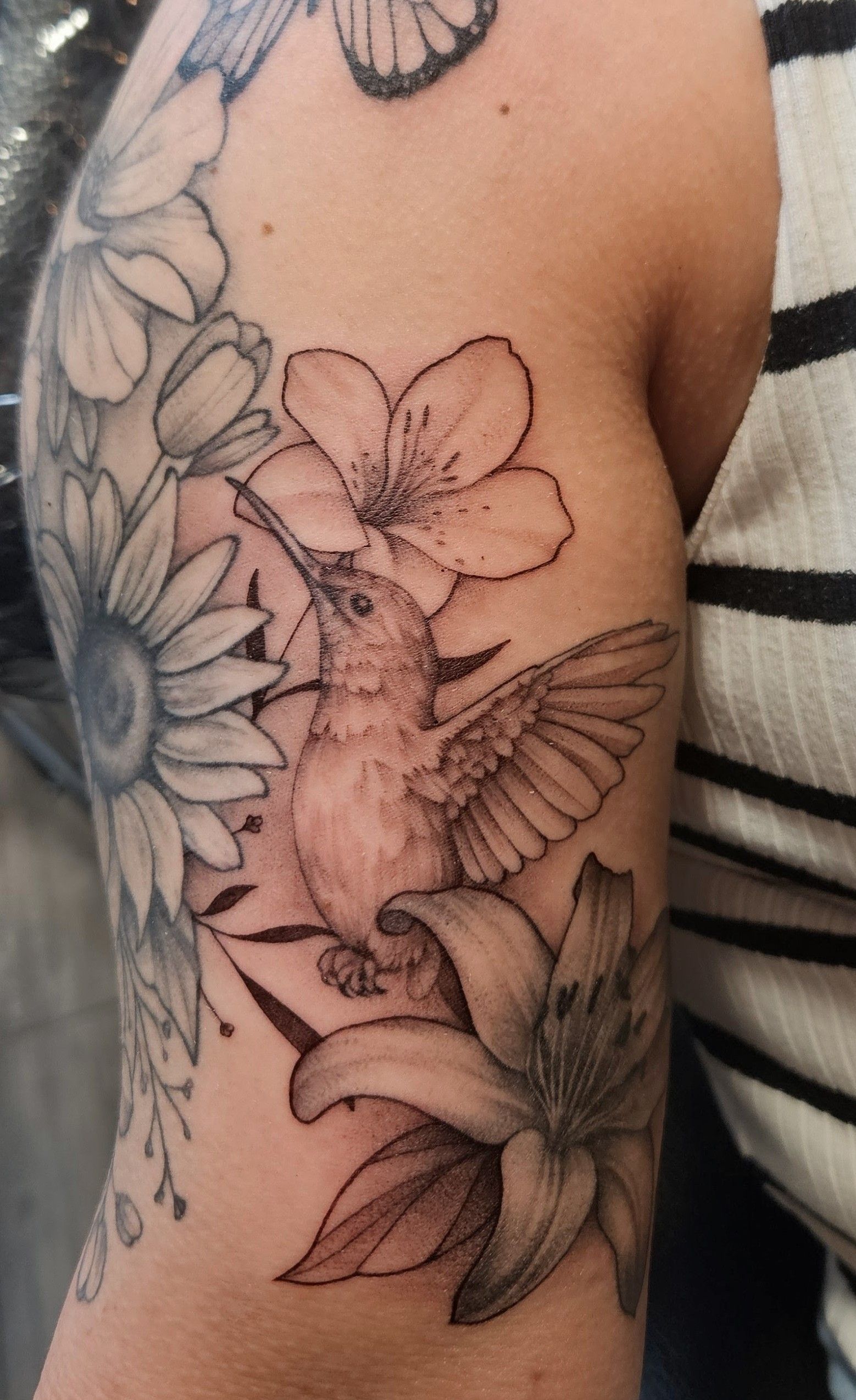 Black-and-white floral tattoo on a person's upper arm with a hummingbird and blossoms