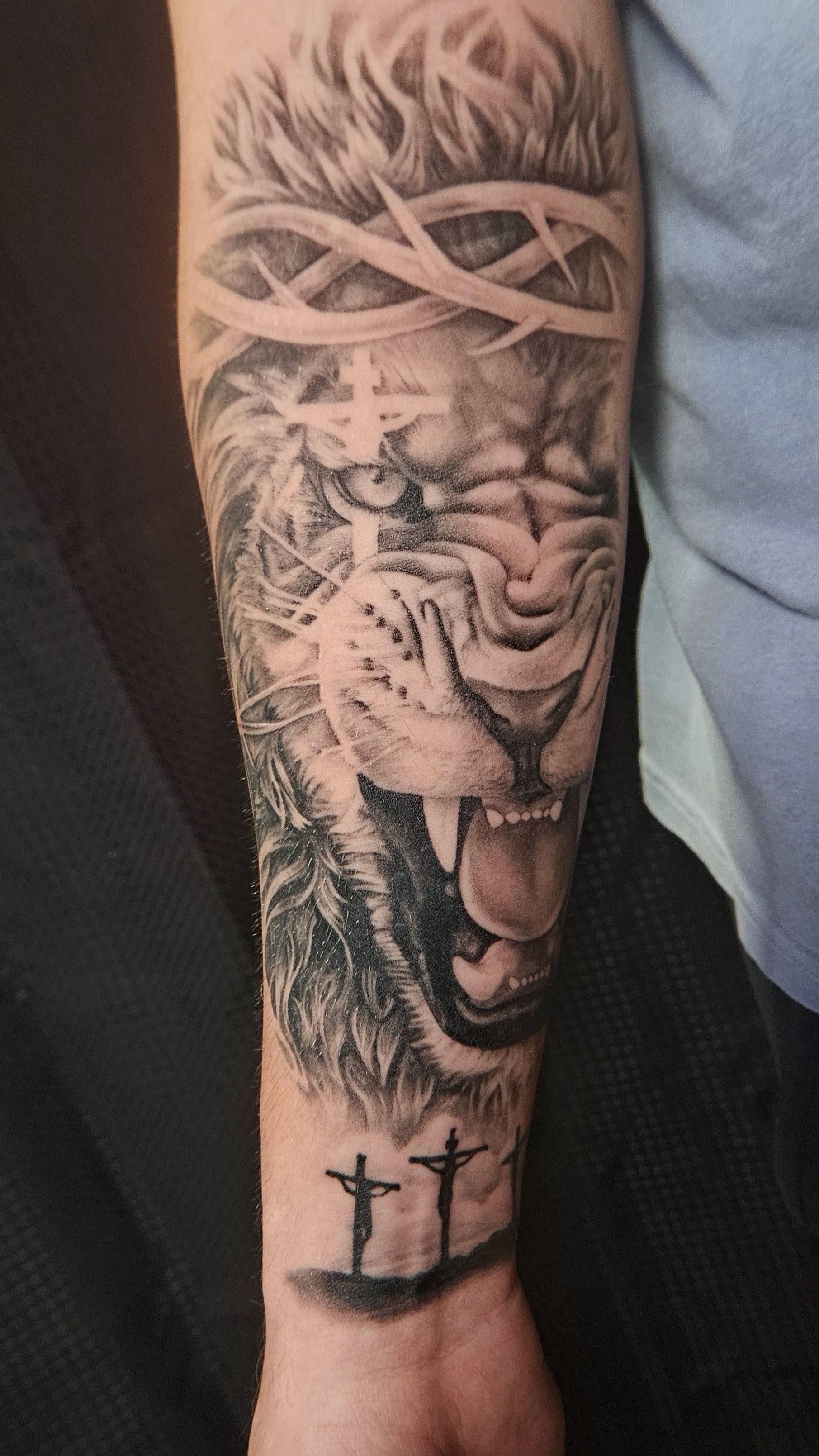 Black-and-gray forearm tattoo of a roaring lion with geometric pattern and two small crosses near the wrist