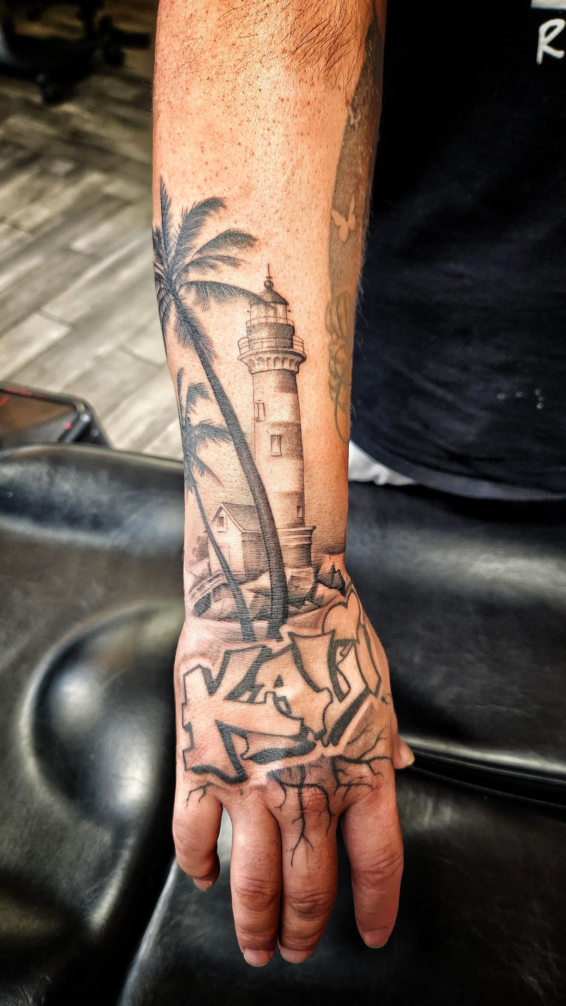 Tattooed forearm and hand resting on a black chair, with a palm tree tattoo visible.