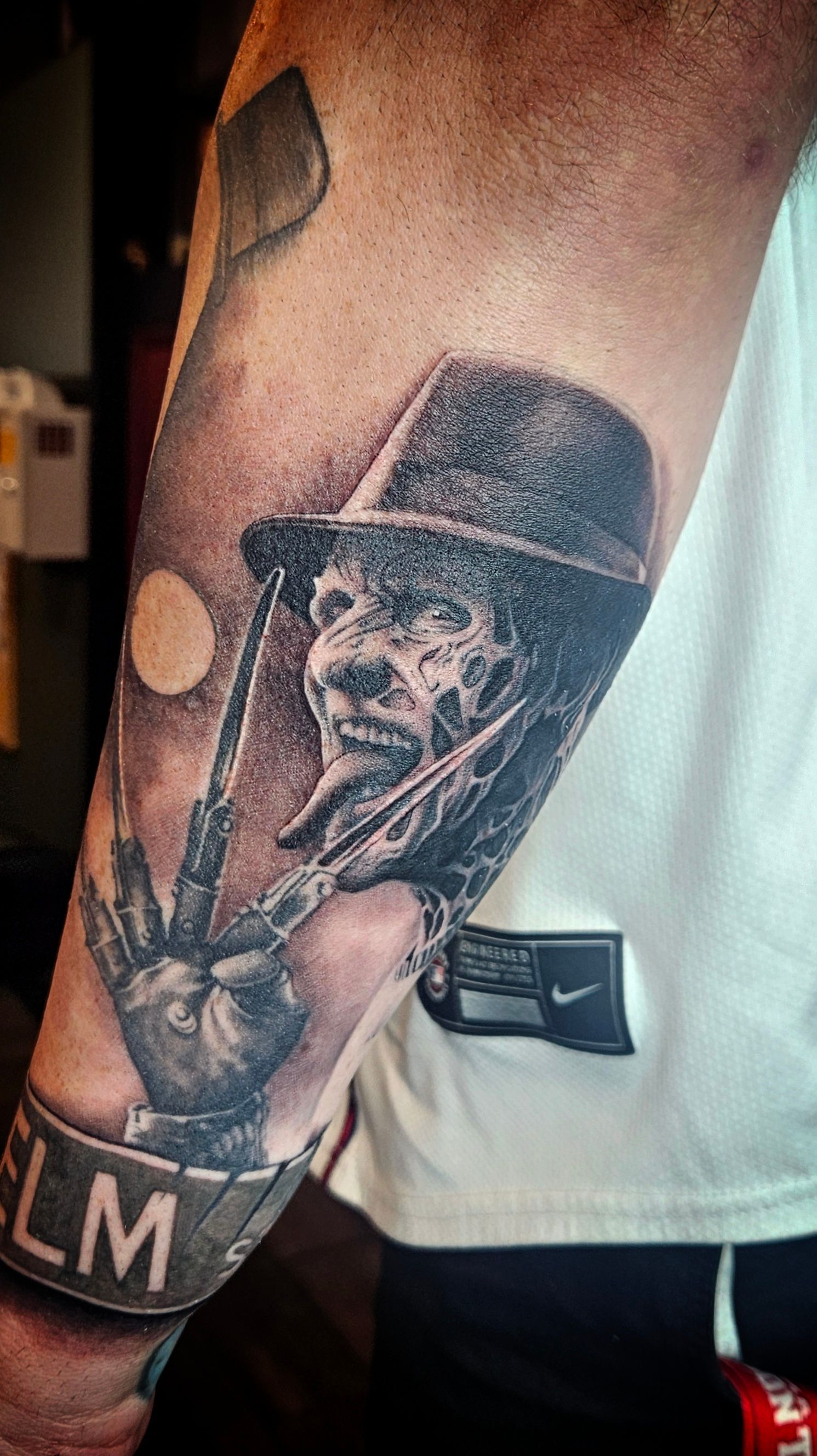Close-up of a forearm tattoo showing a creepy figure in a black hat, with a bracelet and white shirt visible.