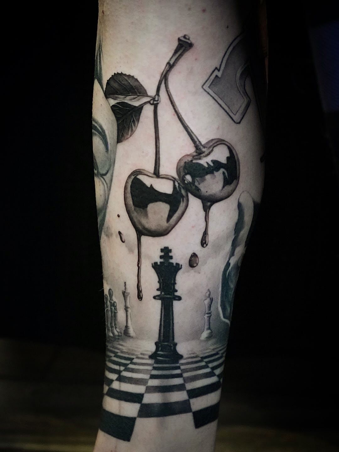 Black-and-white tattoo of a chessboard path with a king piece and hanging cherries on a leg
