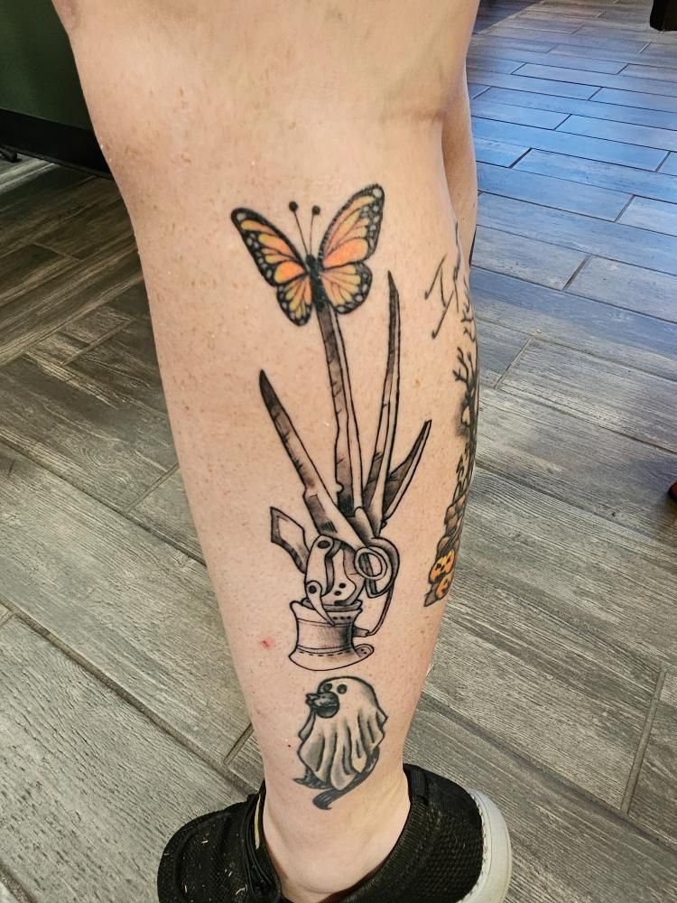 A person has a tattoo of a butterfly , scissors , and a heart on their leg.
