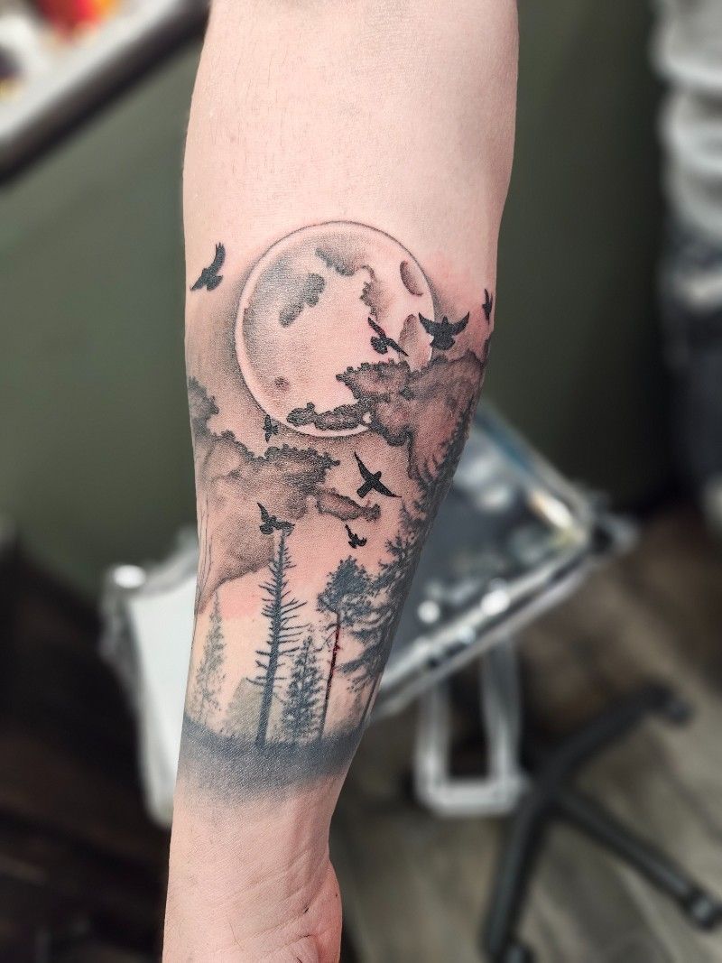 A man has a tattoo of a full moon and birds on his forearm.