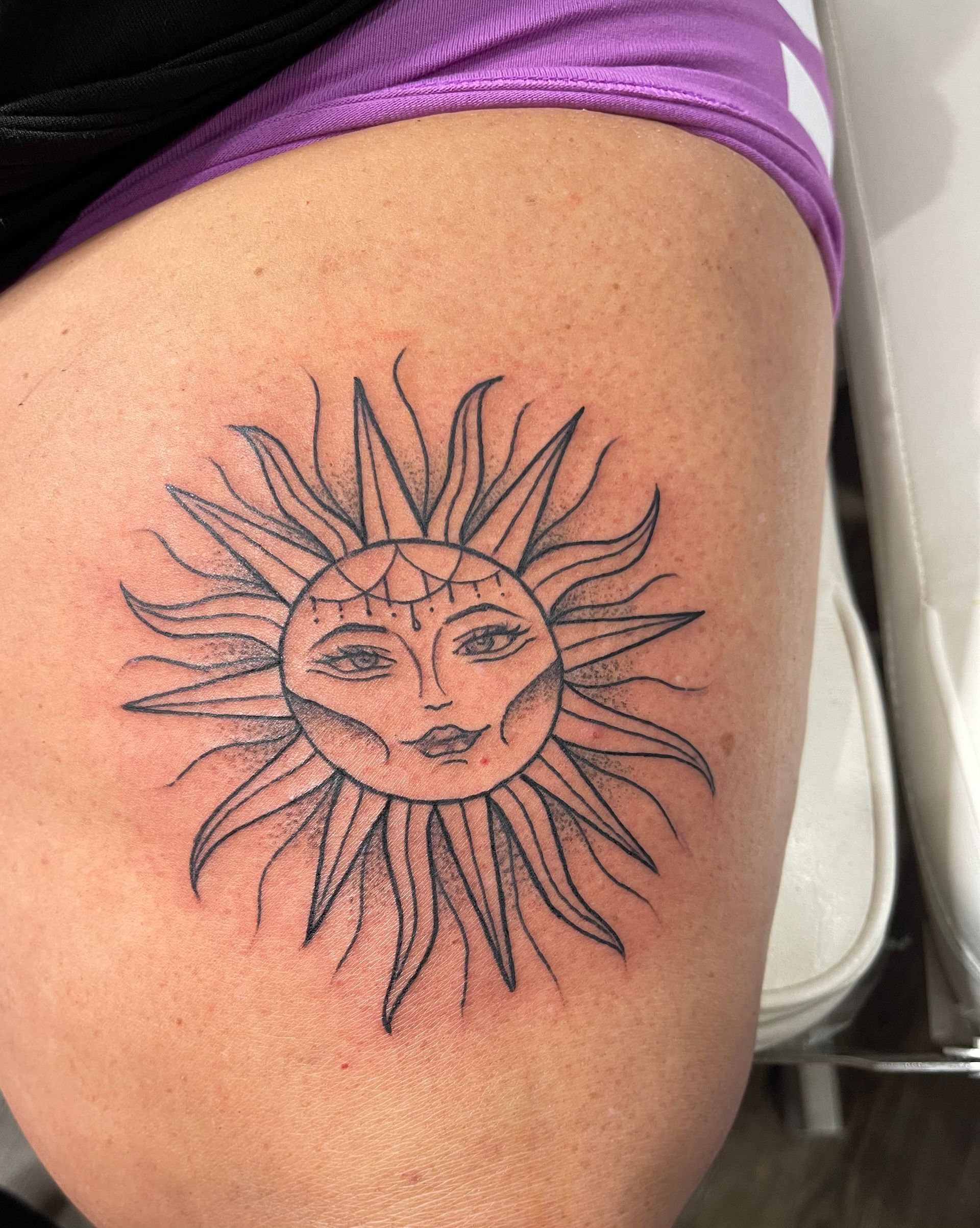 A woman has a tattoo of a sun with a face on her thigh.