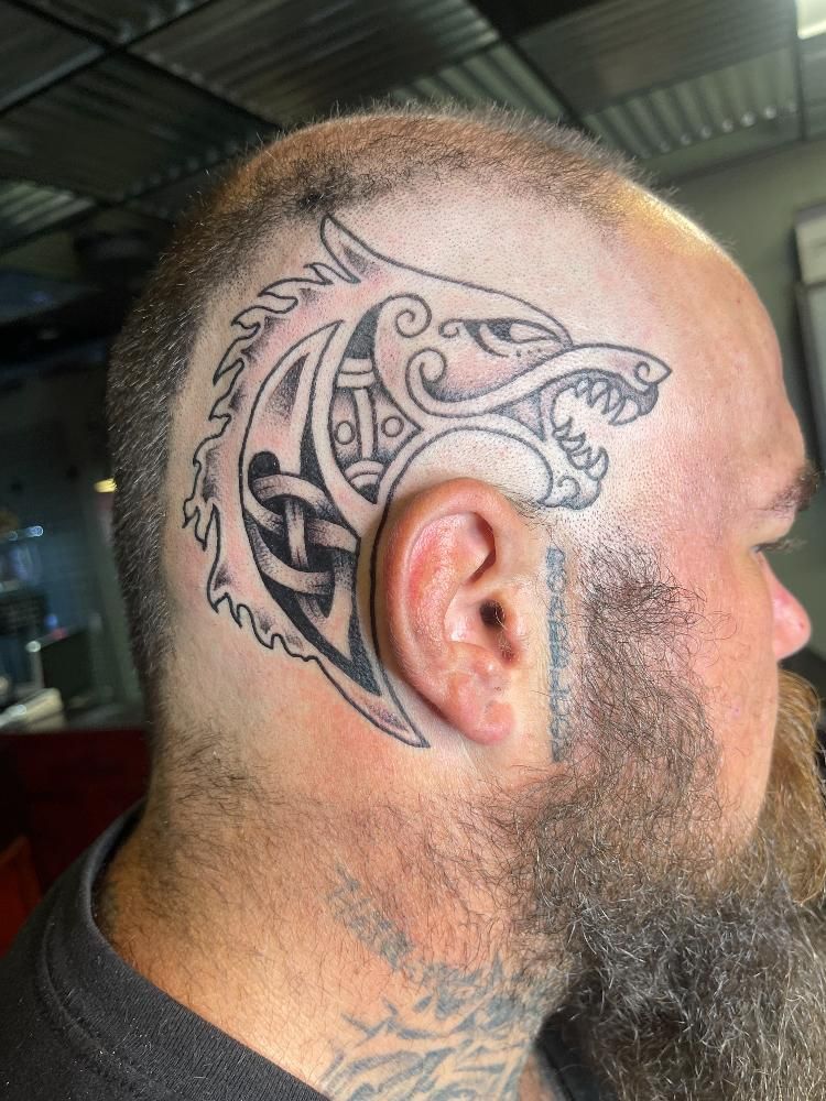 A man with a beard has a tattoo of a wolf on his head.