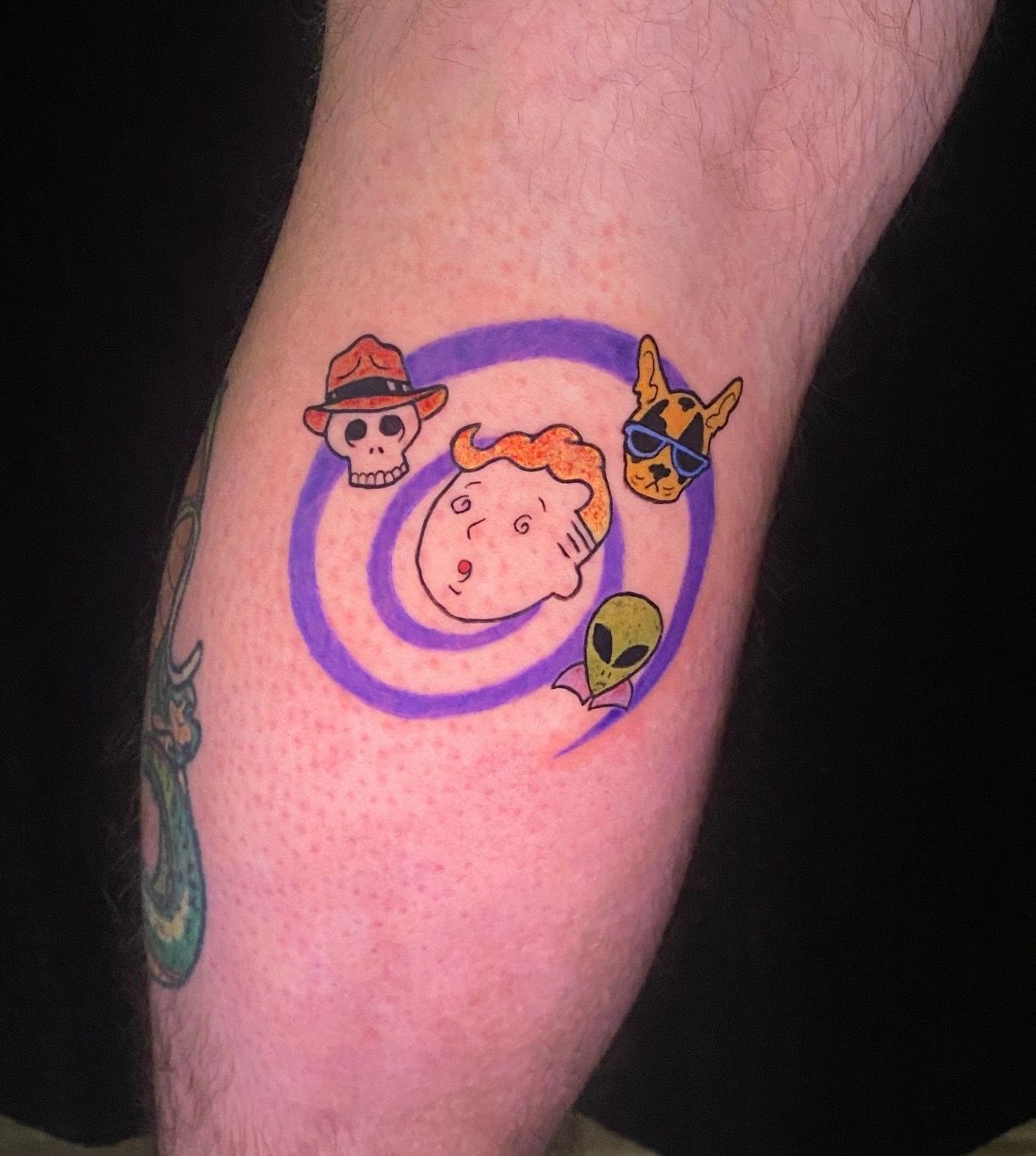 Tattoo on a leg featuring a cartoon face, a skull, a dog, and an alien inside a purple spiral.