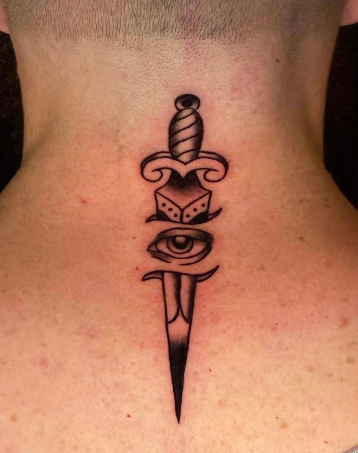 Tattoo on the back of a neck: dagger with a dice, eye, and handle design, black ink.