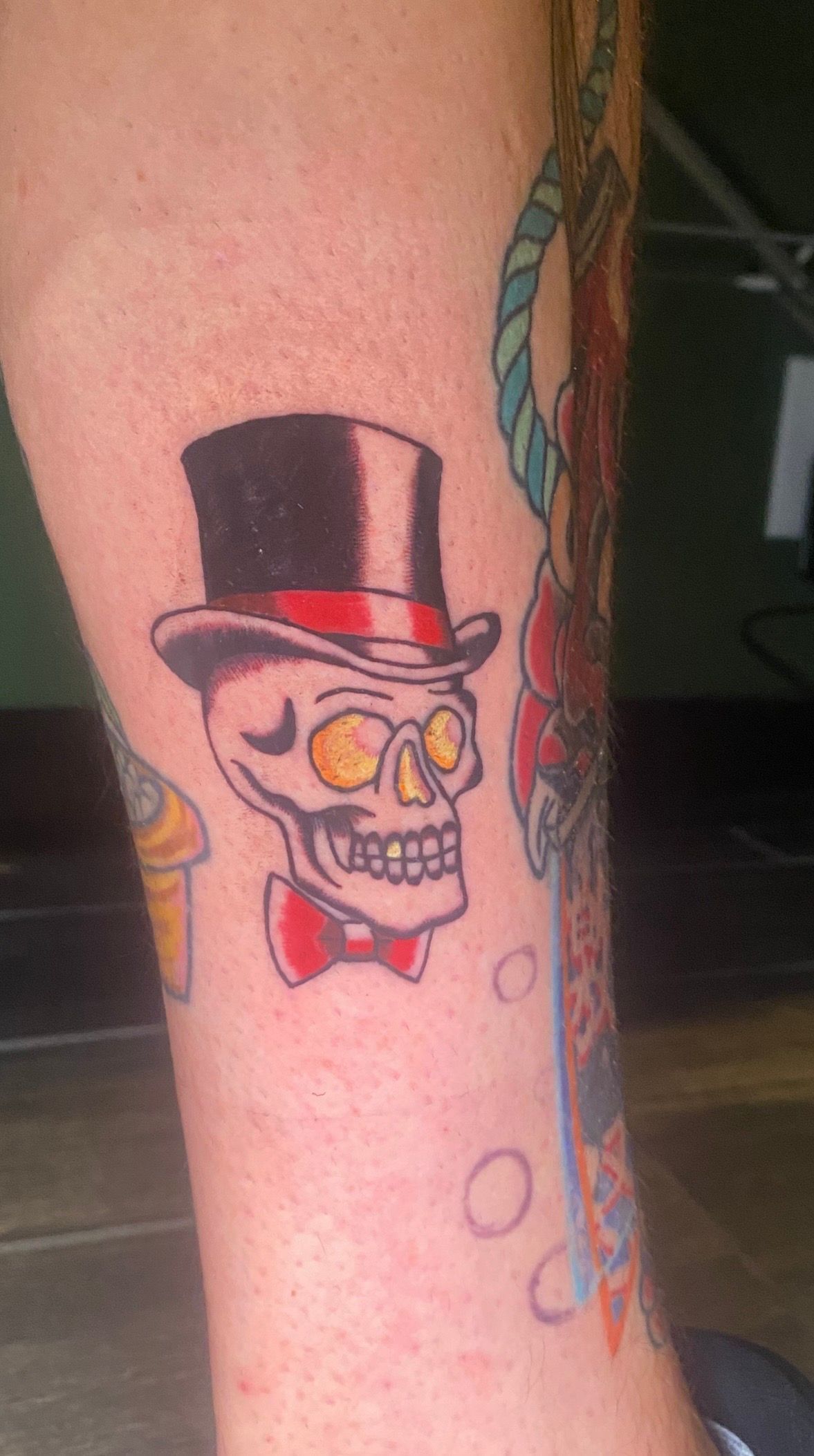 Tattoo of skull with top hat, bow tie, and yellow eyes on a leg.