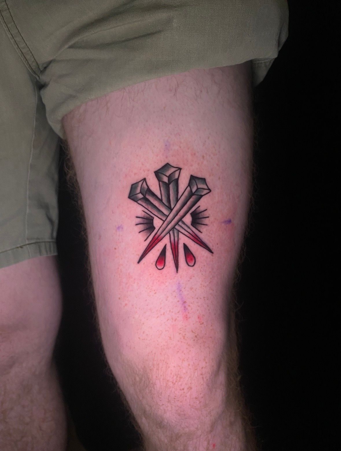 Tattoo of three nails with dripping red blood. On a person's thigh, visible above a khaki shorts leg.