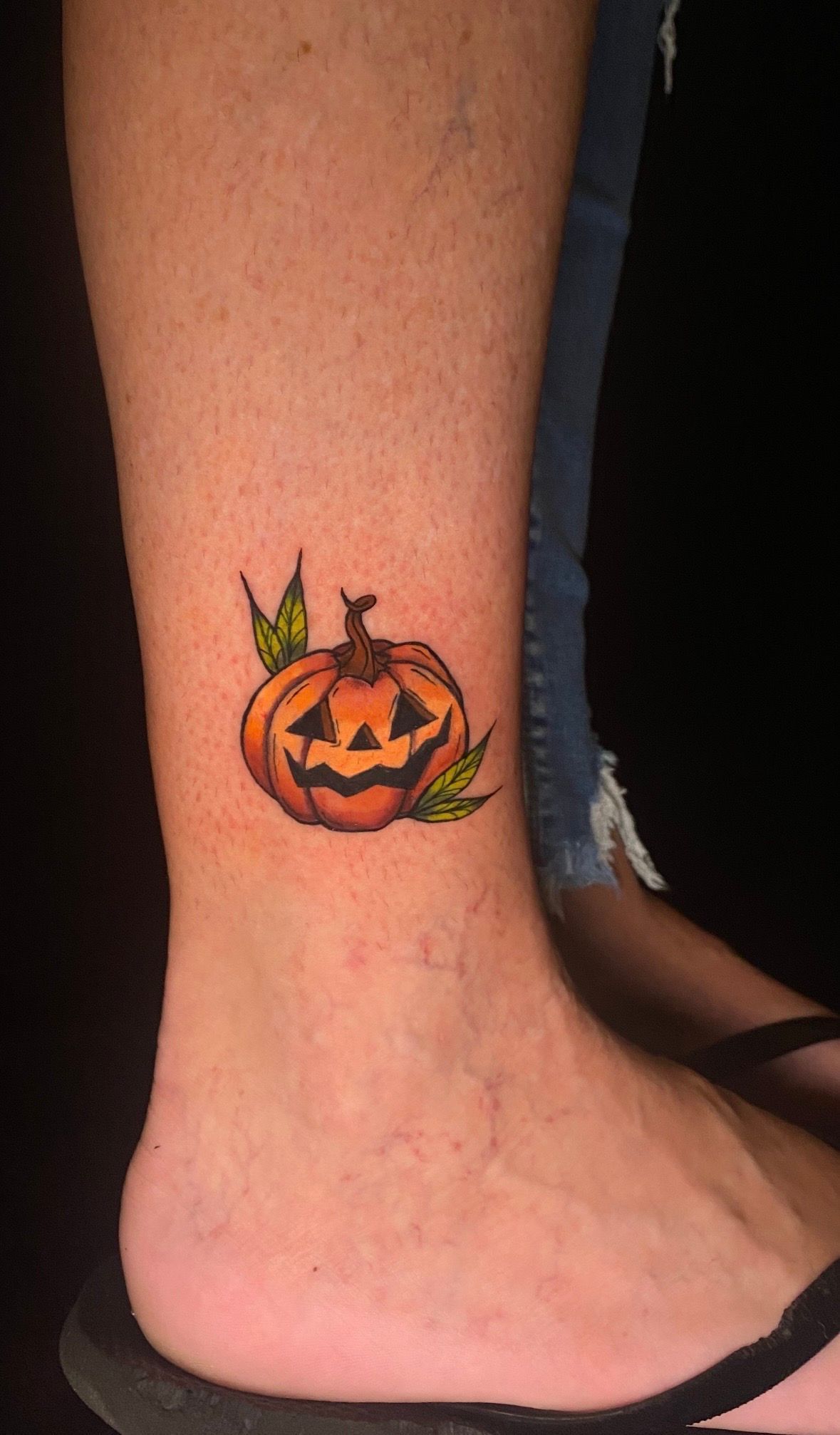 Tattoo of a smiling pumpkin with green leaves on a person's lower leg near the ankle.