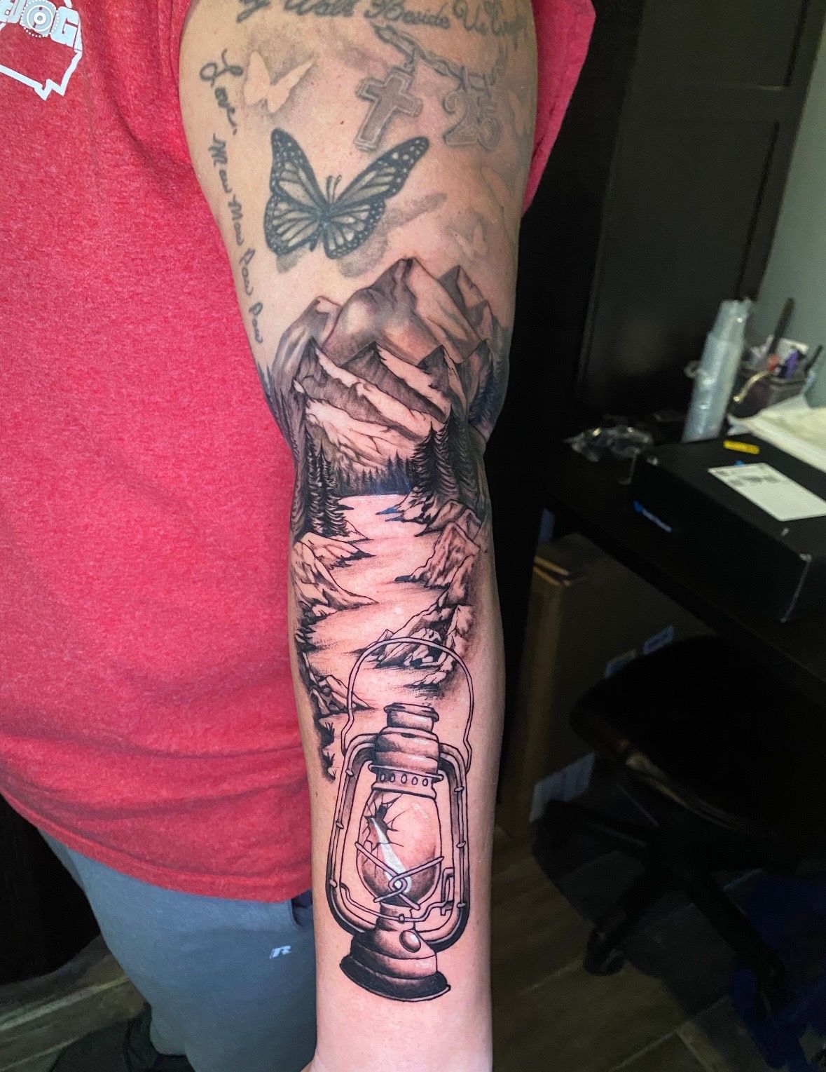 Tattoo on arm depicts mountains, river, butterfly, and lantern in black ink. The arm is on person wearing a red shirt.