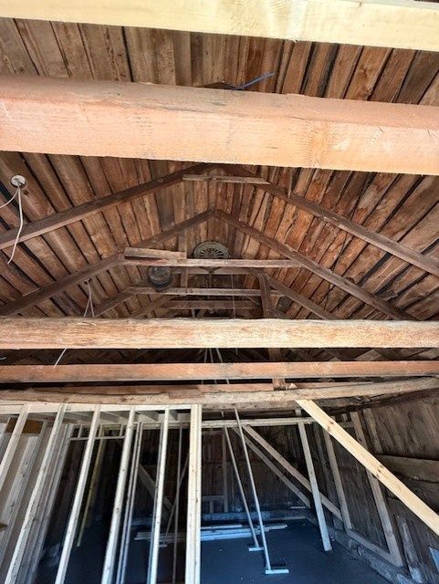 An unfinished attic space with exposed wooden rafters, ceiling beams, and a new wall frame being built.