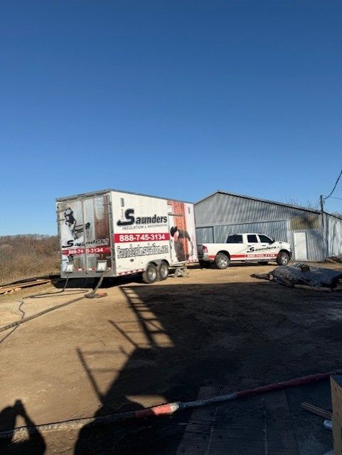A Saunders Moving trailer and white pickup truck parked on a dirt lot in front of a metal building under a clear blue sky.