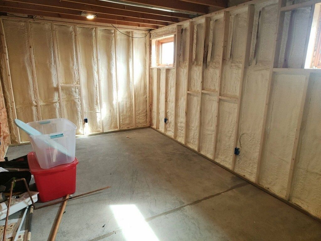 An unfinished basement room with foam-insulated wall studs, concrete floors, and a red storage bin in the corner.