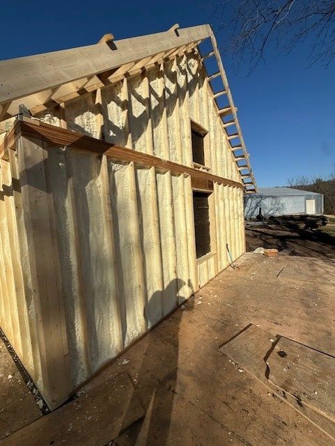 A wooden building under construction with spray foam insulation between wall studs and an open roof frame under blue skies.
