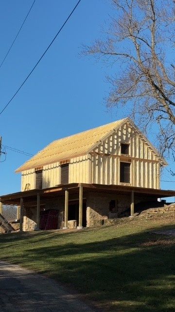 A partially constructed wooden house frame sitting atop a stone foundation on a grassy hillside under a clear blue sky.