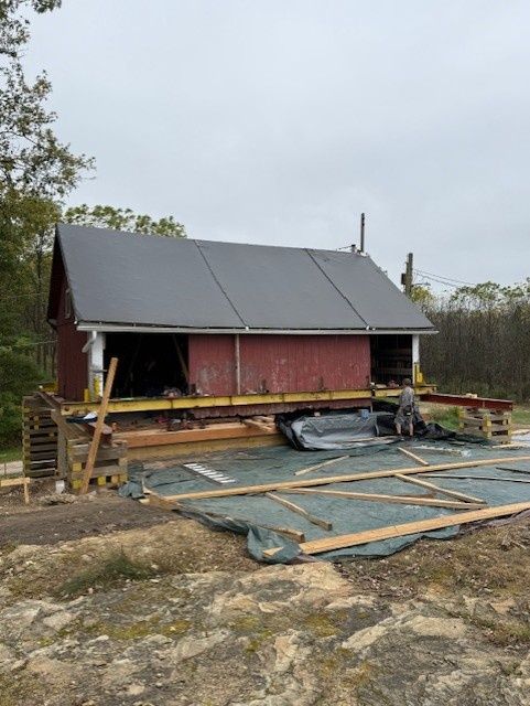 A small red building on a temporary metal frame and wooden cribbing, situated on a dirt lot undergoing construction.