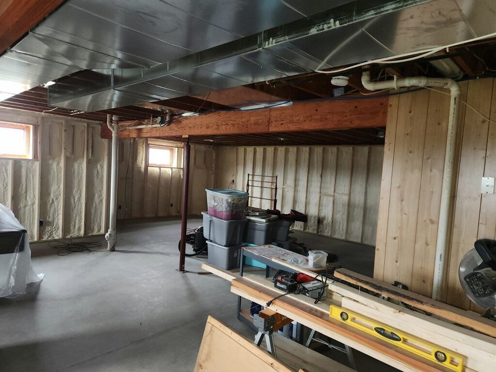An unfinished basement interior with concrete floors, exposed ceiling ducts, framed walls with insulation, and work tools.