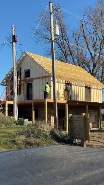 Two workers in high-visibility gear frame the upper level of a two-story residential building under a clear blue sky.