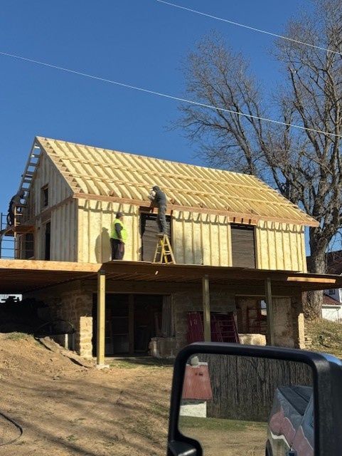 Two workers renovate a wooden framed building with an exposed stone foundation under a clear blue sky.