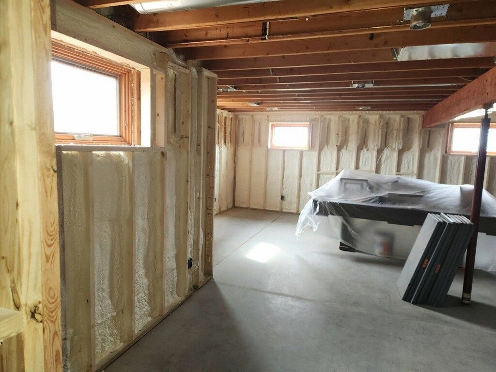 An unfinished basement with exposed wooden beams, foam-insulated walls, concrete floors, and two small windows.