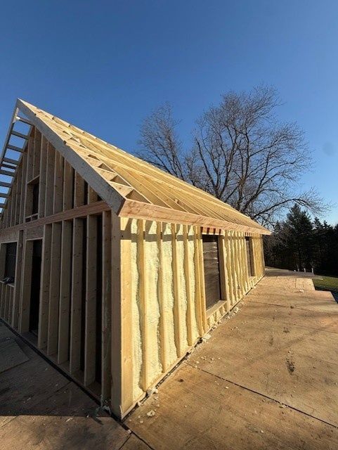 A wood-framed building under construction with yellow spray foam insulation between studs under a clear blue sky.