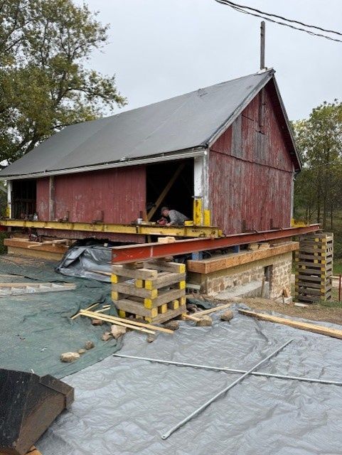 A red, rustic barn elevated on steel beams and timber cribbing stacks for structural repair above a stone foundation.
