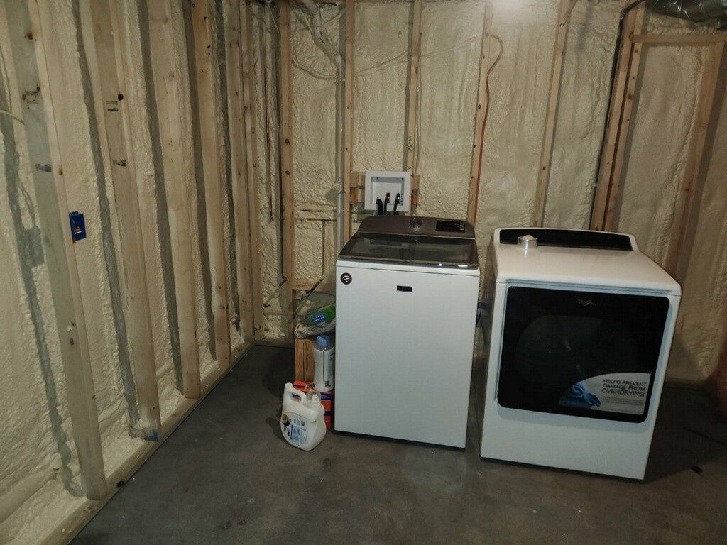 A washing machine and dryer sit against unfinished basement walls with spray foam insulation and exposed wooden studs.