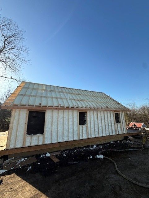 A framed, insulated house structure under a clear blue sky, showing wooden studs and white foam insulation.