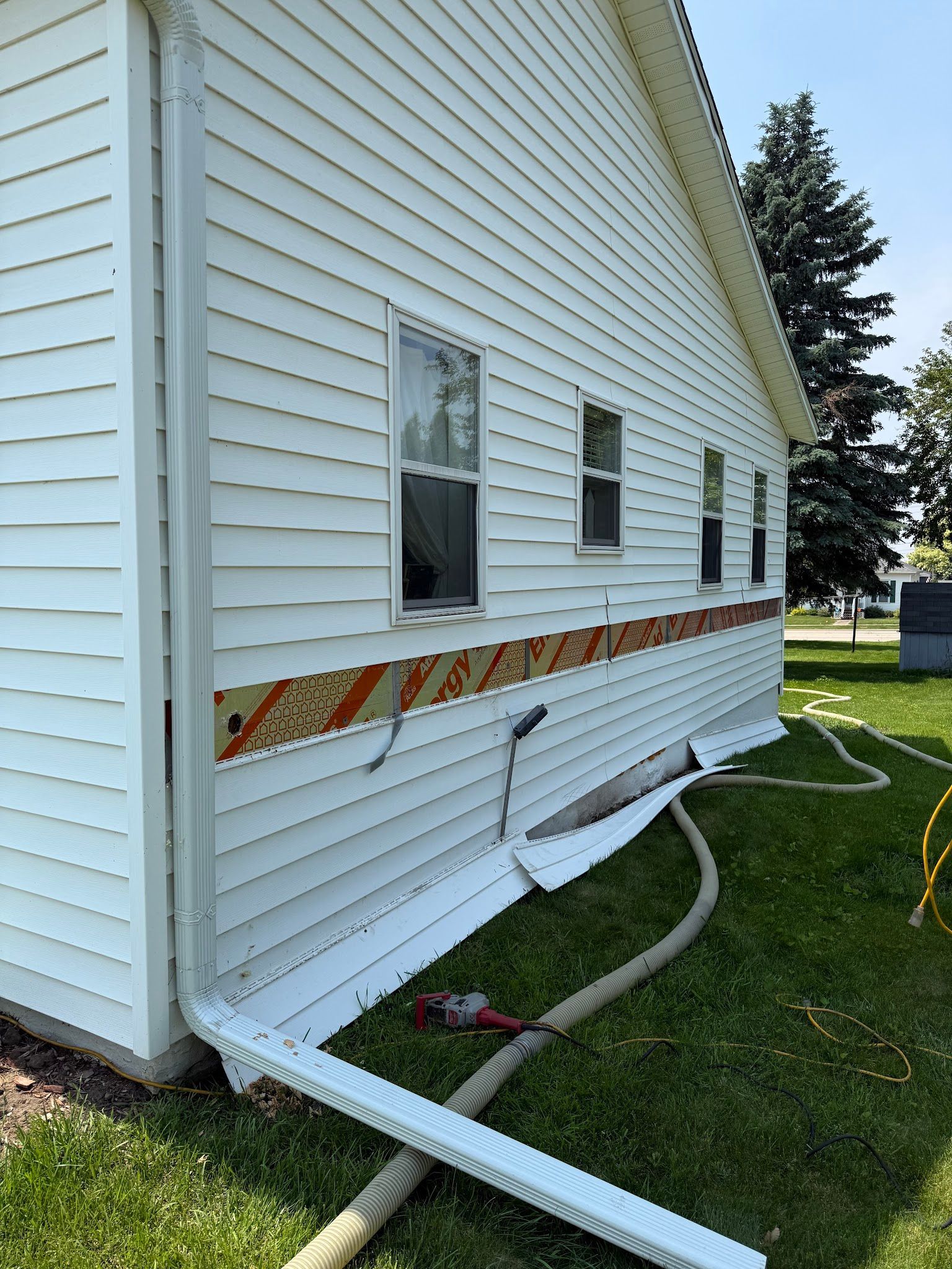 The side of a white-sided house under repair, with a strip of sheathing visible and a white downspout lying on the grass.