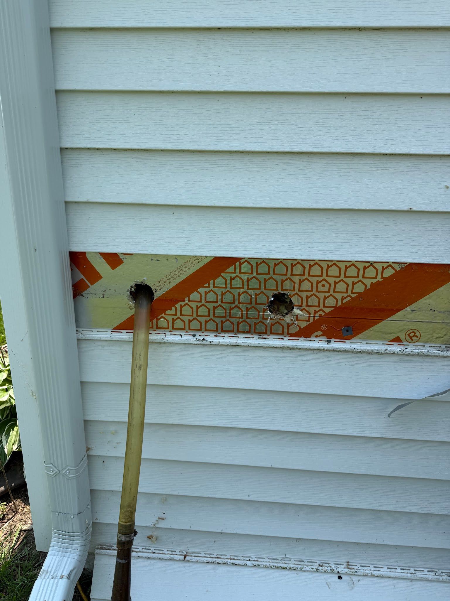 A section of white house siding is missing, revealing orange insulation foam and a pipe exiting a hole in the wall.