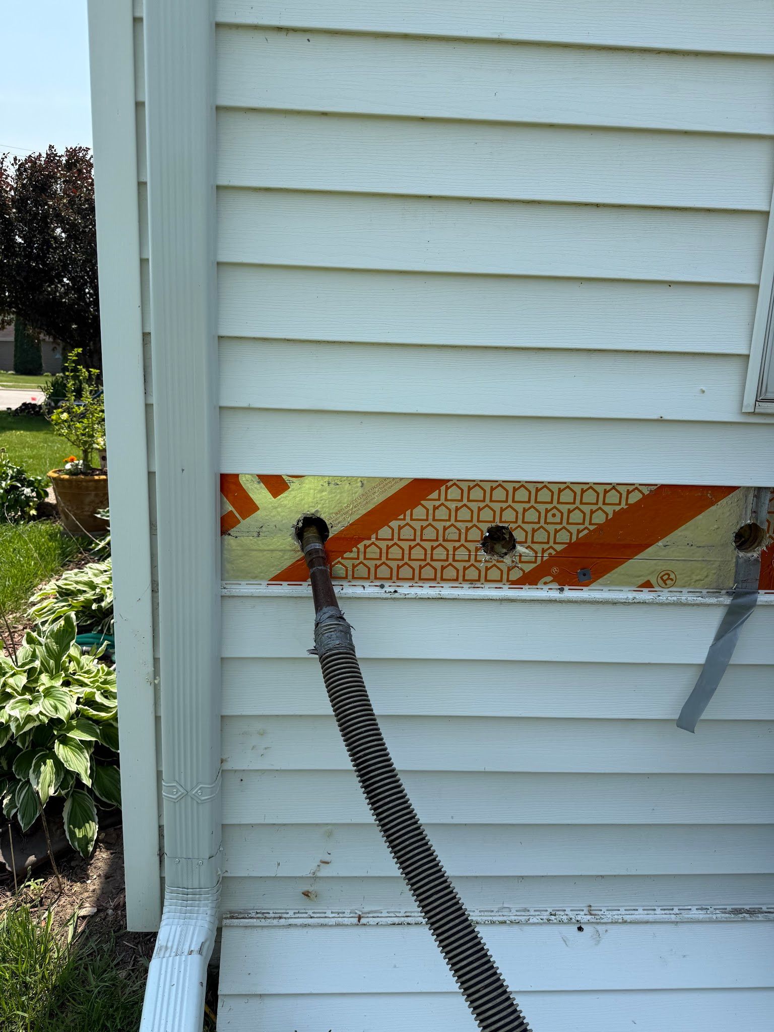 A hole in white siding reveals orange insulation, with an electrical cable hanging out and a hanging strip of gray tape.