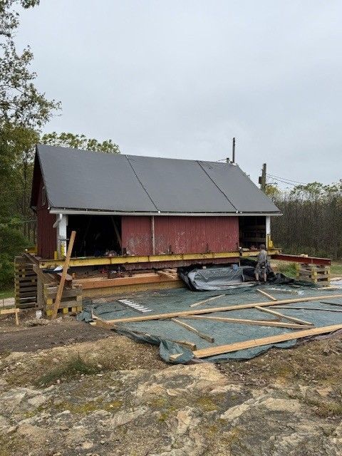 Red barn under construction with gray roof panels and lumber scattered on rocky ground