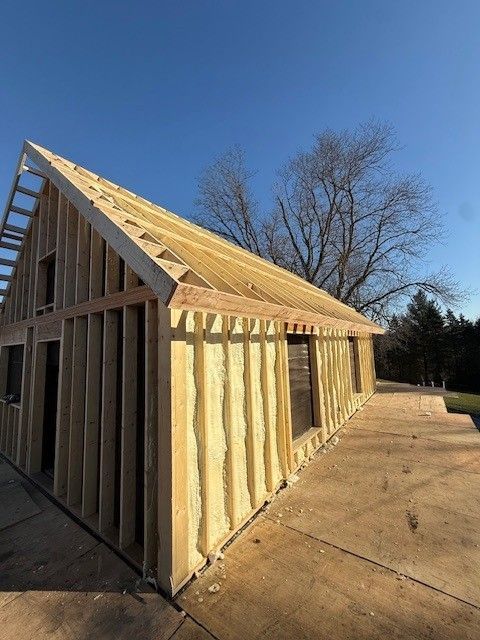 Wood-framed house under construction with exposed studs and roof trusses on a concrete slab under a blue sky