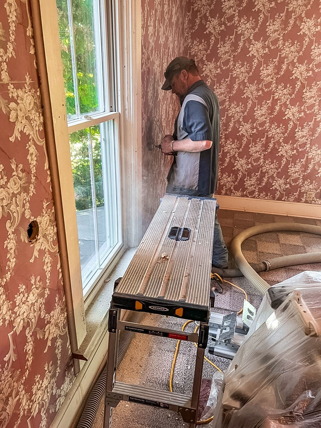 A man is standing on a ladder in a room next to a window.