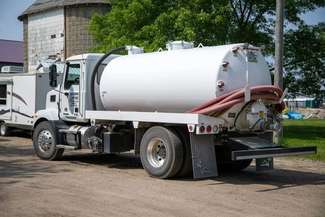 White septic truck on gravel road with a trailer, near a barn and trees.