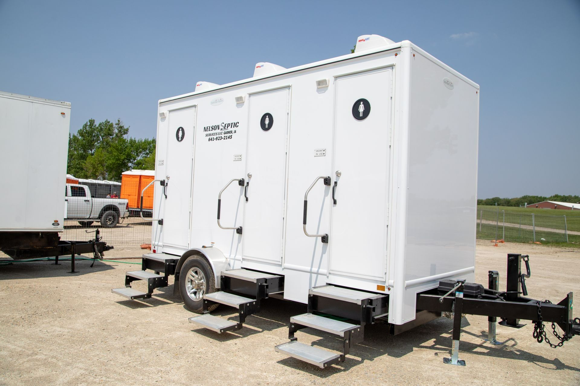 White portable restroom trailer with three stalls, parked outdoors.