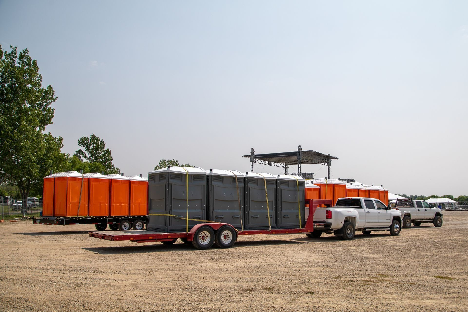 Portable toilets on trailers, being transported by trucks, on a dirt lot. Orange and gray units visible.