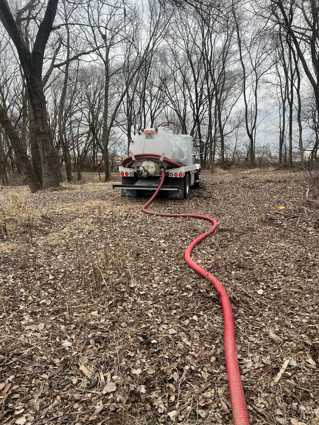 Water truck in a brown, leaf-covered field, red hose extending away, trees in background on a cloudy day.