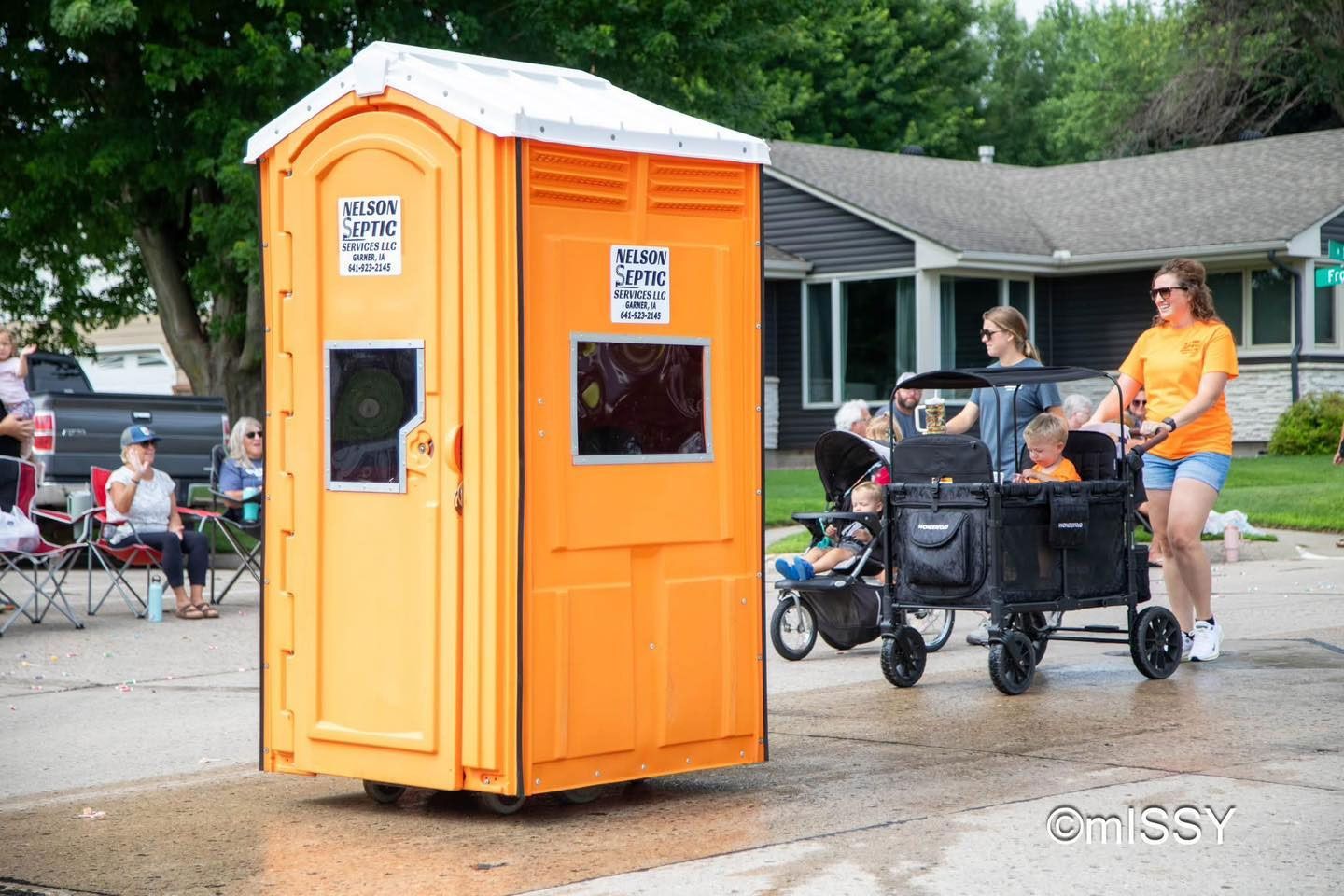 Orange portable toilet rolls down a street during a parade, with people watching from the sidewalk and walking beside it.