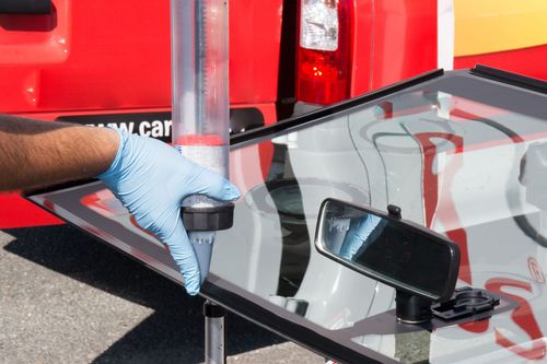 Gloved hand using a suction cup to repair a cracked car windshield near a red vehicle
