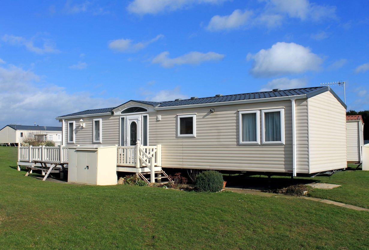 A beige mobile home with a small wooden deck sits on a grassy field under a sunny blue sky.