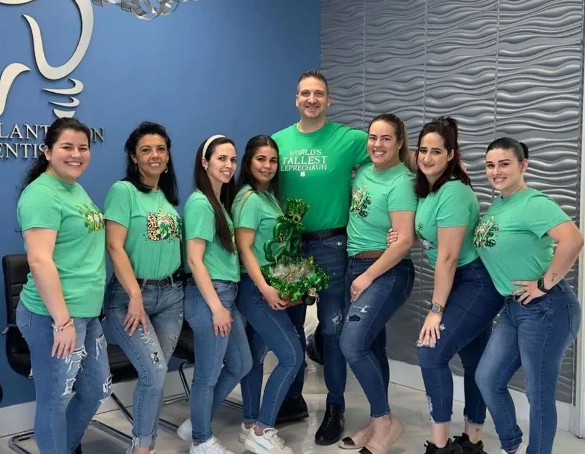 Dental office staff in green shirts and jeans pose near a blue wall, celebrating St. Patrick's Day.